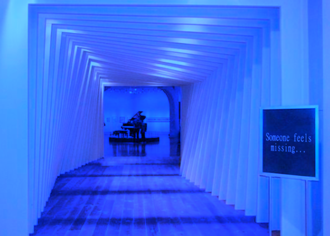 A twisting hallway, drenched in blue light, leads the way to an empty room with a grand piano. On the right of the entrance, a sign reads: "Someone feels missing..."